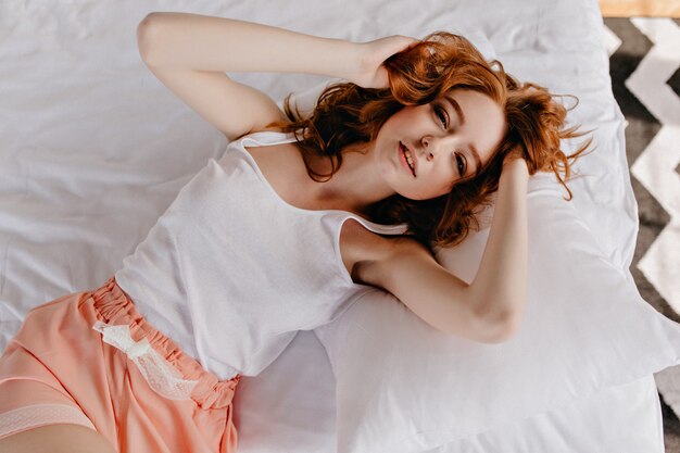 Blithesome femme allongée sur un lit moelleux. Vue aérienne d'une superbe fille au gingembre avec une coiffure frisée.