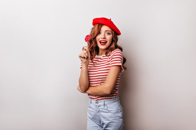 Blithesome femme bouclée mangeant une sucette sur un mur blanc. Charmante fille française en béret posant avec des bonbons.