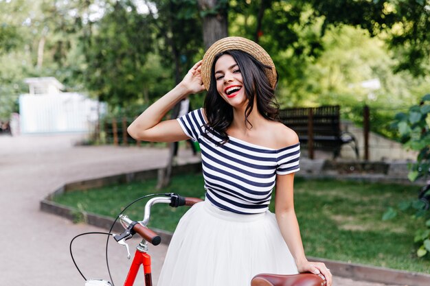 Blithesome fille caucasienne au chapeau posant sur la nature. Superbe femme brune en jupe debout dans la rue à vélo.