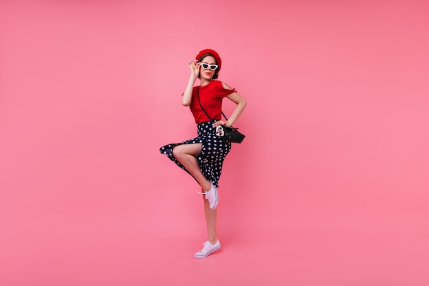 Blithesome fille mince dans la danse du béret français. Belle dame aux cheveux courts en jupe noire sautant sur un mur rose.