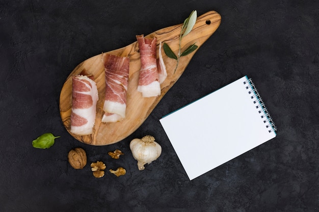 Bloc-notes blanc à spirale avec bacon roulé sur une planche en bois au basilic; club de noix et d&#39;ail