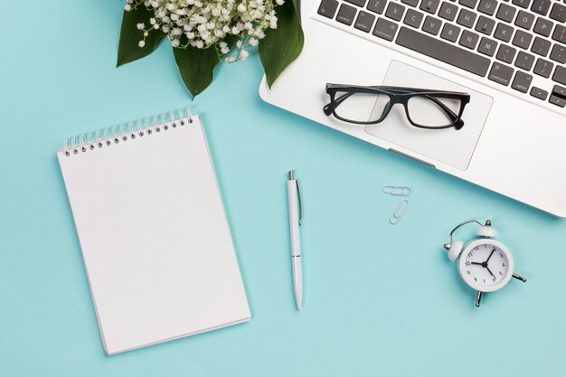 Bloc-notes à spirale, stylo, trombone, réveil, lunettes de vue sur ordinateur portable avec bouquet de muguet sur un bureau bleu