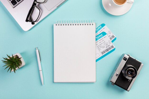 Bloc-notes en spirale vierge avec billets d&#39;avion entourée d&#39;un ordinateur portable, lunettes de vue, stylo, appareil photo, tasse à café sur le bureau bleu