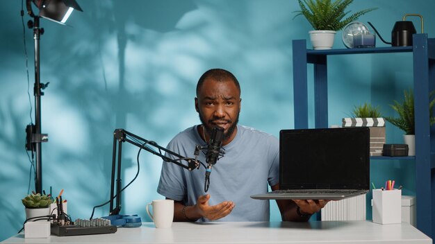 Blogueur afro-américain faisant une revue de produit d'un ordinateur portable à la caméra pendant l'enregistrement pour la chaîne vlog. Un vlogger noir passe en revue le gadget en vidéo pour un podcast sur les réseaux sociaux. Homme avec appareil
