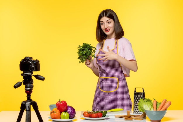 Blogueur culinaire jeune fille en tablier rose enregistrant une vidéo pour les médias sociaux tenant un persil