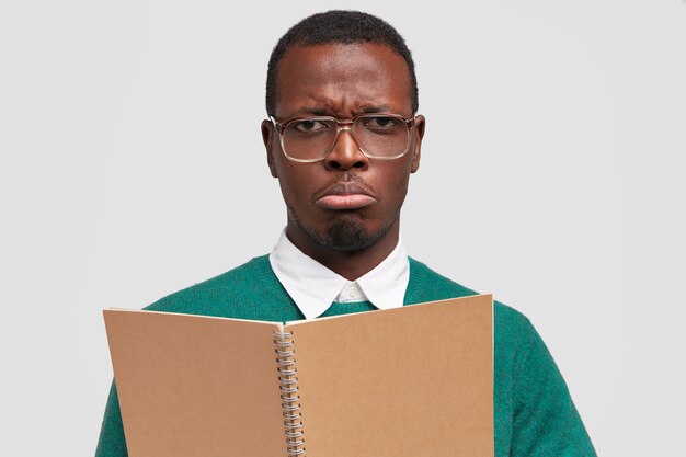 Un blogueur homme triste prend des notes pour publication, porte un bloc-notes, porte de grandes lunettes maladroites, porte un sac à main, a déplu à l'expression du visage