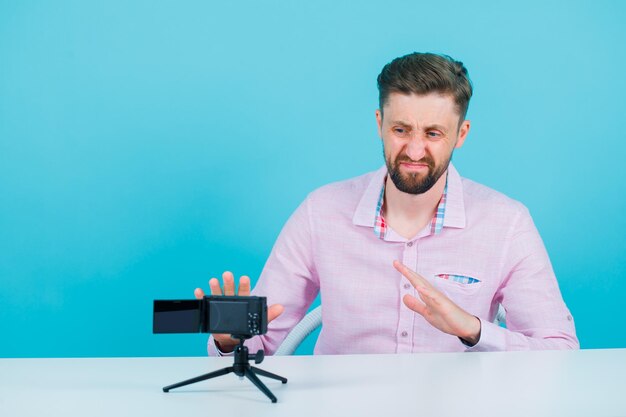 Un blogueur insatisfait pose devant sa mini caméra en s'asseyant sur fond bleu