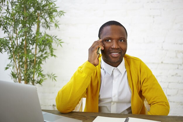 Blogueur masculin à la peau sombre à la mode ayant une conversation téléphonique, assis devant un ordinateur portable ouvert, travaillant sur le contenu de son blog de voyage. Personnes, travail, profession et gadgets électroniques modernes