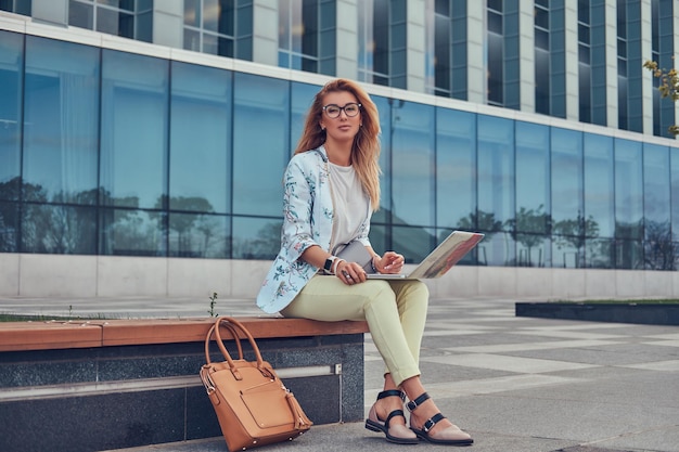 Blogueur de mode élégant se relaxant en plein air, travaillant sur l'ordinateur portable, assis sur un banc contre un gratte-ciel.