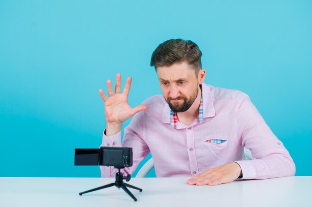 Un blogueur pose devant sa mini caméra en montrant un geste de griffes sur fond bleu