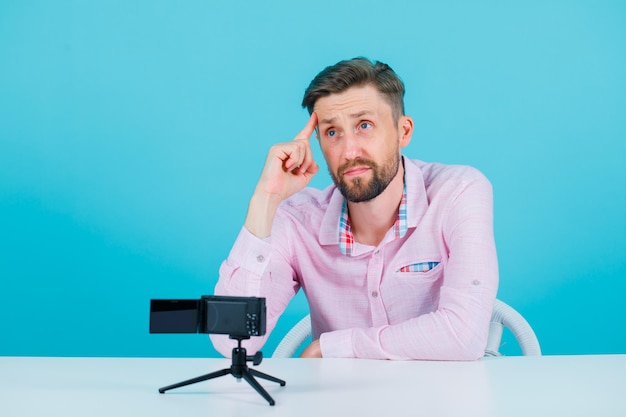 Un blogueur pose devant sa mini caméra en tenant l'index sur la tempe sur fond bleu