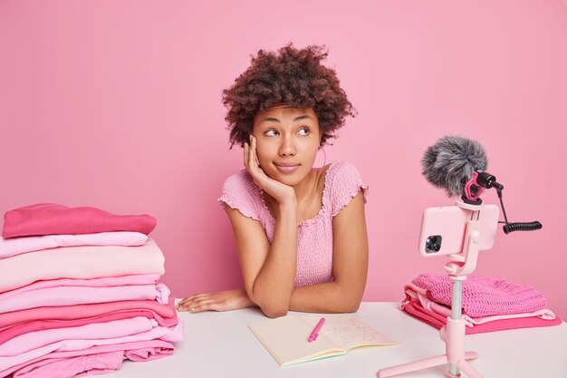 Une blogueuse afro-américaine pensive enregistre un didacticiel vidéo sur la façon de se laver correctement, assis à table avec des piles de cahiers ouverts de linge plié isolés sur du rose