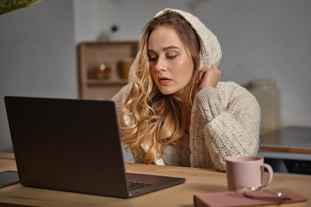 Blogueuse blonde en sweat à capuche beige assise dans la cuisine à une table en bois regardant son livre d'ordinateur portable ...