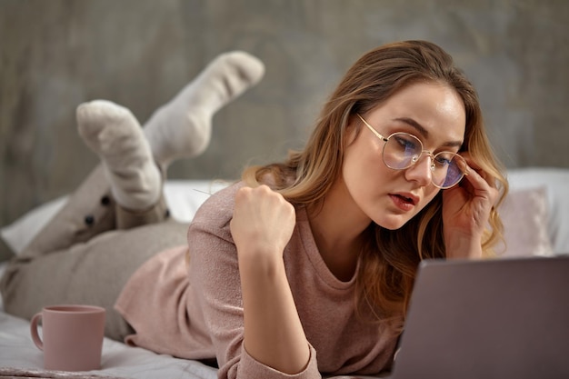 Blogueuse blonde en vêtements décontractés à lunettes allongée en tailleur sur le lit avec une tasse en regardant sa la...