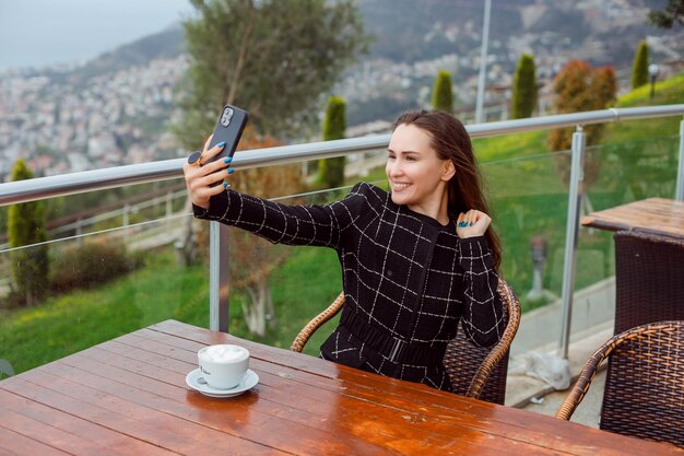 Une blogueuse heureuse prend un selfie avec son smartphone en levant la main en s'asseyant sur fond de vue sur la ville