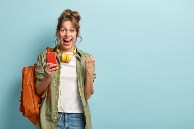 Une blogueuse heureuse serre le poing, se sent excitée par les statistiques sur la page Web des réseaux sociaux, utilise un téléphone portable et des écouteurs, porte une chemise et un jean kaki, porte un sac à dos, isolé sur un mur bleu