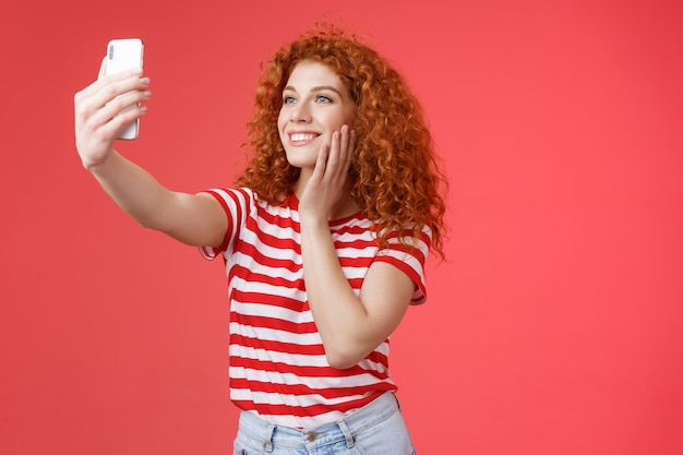 Une blogueuse de mode de vie populaire élégante des médias sociaux adore prendre des photos elle-même étendre le bras tenir le smartphone posant des vêtements d'été mignons et idiots faire selfie souriant écran de téléphone tendre fond rouge.