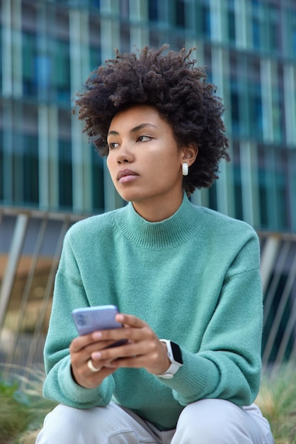 Une blogueuse pensive discute avec des abonnés sur les réseaux sociaux sur un smartphone moderne surfe sur les réseaux sociaux vêtue de vêtements décontractés à l'extérieur