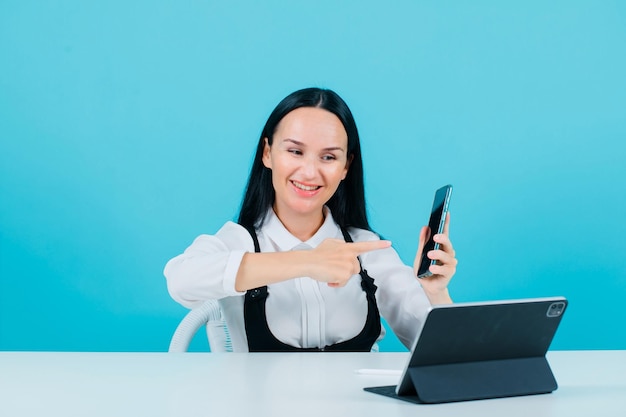 Une blogueuse souriante montre son téléphone avec l'index en s'asseyant devant la caméra de la tablette sur fond bleu