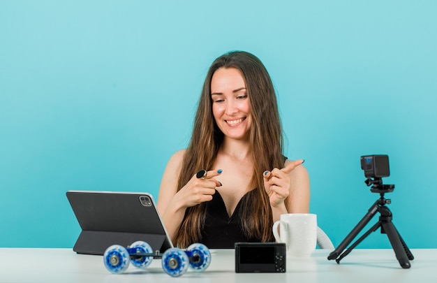 Une blogueuse souriante pointe vers la droite avec les index en regardant l'écran de la tablette sur fond bleu