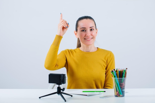 Une blogueuse souriante pointe vers le haut avec l'index sur fond blanc