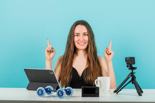 Une blogueuse souriante pointe vers le haut avec l'index en regardant la caméra sur fond bleu