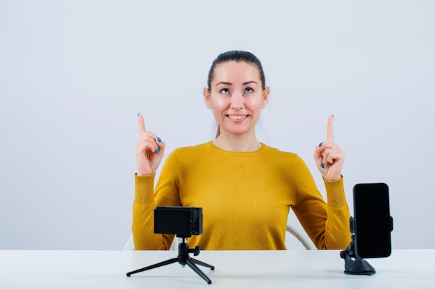 Une blogueuse souriante pointe vers le haut avec l'index en s'asseyant devant sa mini caméra sur fond blanc