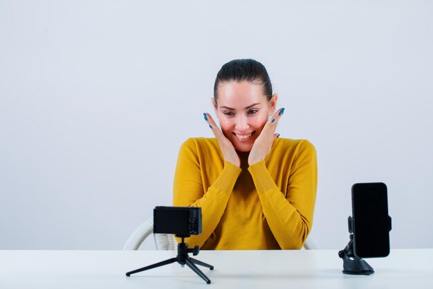 Une blogueuse souriante pose en se tenant la main près des joues devant sa mini caméra sur fond blanc