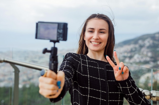 Une blogueuse souriante prend un selfie en montrant un geste de victoire sur fond de vue sur la ville
