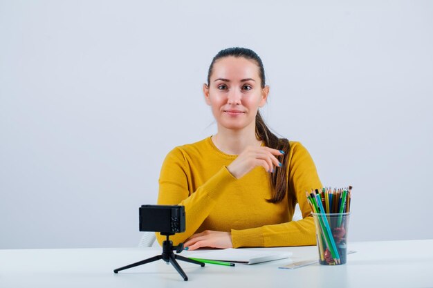 Une blogueuse souriante regarde loin en s'asseyant devant sa mini caméra sur fond blanc