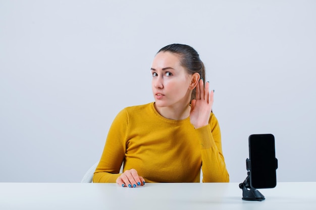 Une blogueuse tient la main derrière l'oreille en s'asseyant devant une caméra mobile sur fond blanc