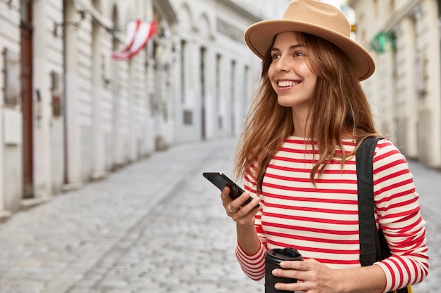 Une blogueuse de voyage heureuse utilise une application d'itinéraire, tient un téléphone intelligent moderne, se promène dans une ville ancienne, boit du café à emporter