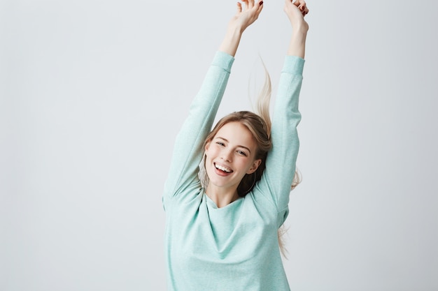 Blonde belle jeune femme en haut bleu clair étirant les bras de bonne humeur comme pour célébrer la victoire. Femme largement souriante montrant des dents blanches et des émotions positives, s'amusant à l'intérieur.