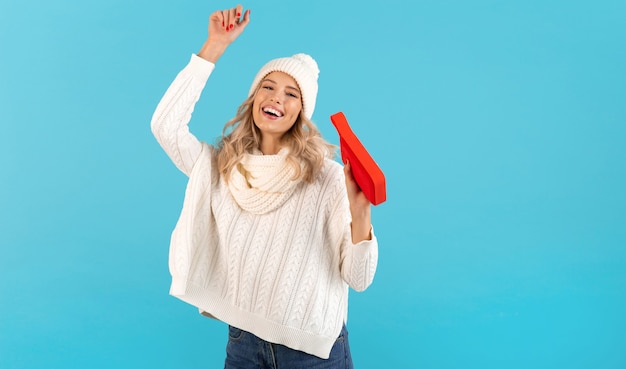 Blonde élégante souriante belle jeune femme tenant un haut-parleur sans fil écoutant de la musique joyeuse danse portant un pull blanc et un chapeau tricoté à la mode de style hiver posant