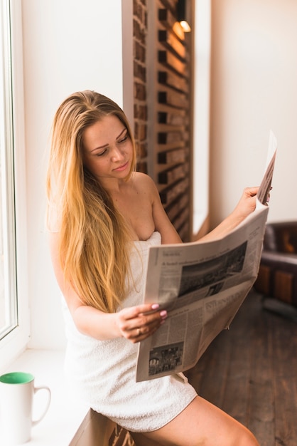 Photo gratuite blonde jeune femme enveloppée sur une serviette en lisant le journal