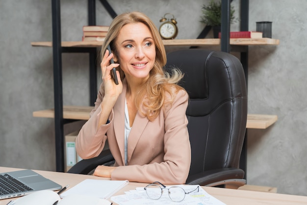 Blonde jeune femme parlant au téléphone assis sur une chaise au bureau