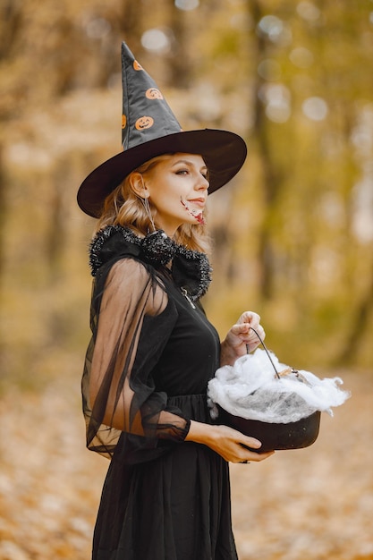 Blonde jeune fille sorcière en forêt à l'Halloween. Fille vêtue d'une robe noire et d'un chapeau conique. Sorcière tenant un truc de magicien.