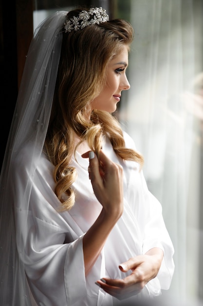 Photo gratuite blonde mariée à la couronne se dresse devant la fenêtre brillante du matin