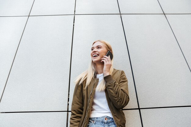 Blonde souriante, parler au téléphone tout en se tenant près du mur