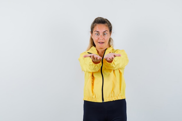 Blonde woman stretching hands comme tenant quelque chose d'imaginaire en blouson aviateur jaune et pantalon noir et à la surprise