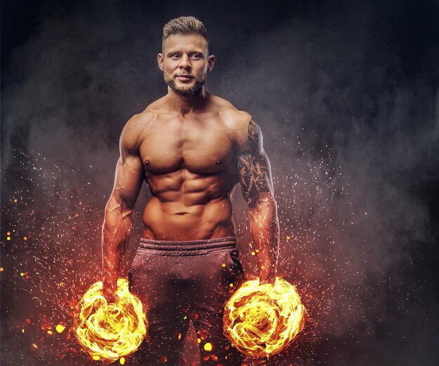 Bodybuilder élégant et puissant avec tatouage sur son bras, posant avec des haltères dans un studio. Concept d'art du feu.