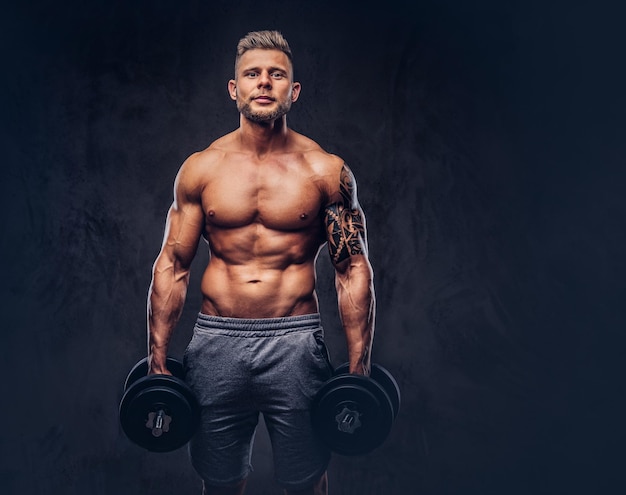 Photo gratuite bodybuilder élégant et puissant avec tatouage sur son bras, posant avec des haltères dans un studio. isolé sur un fond sombre.