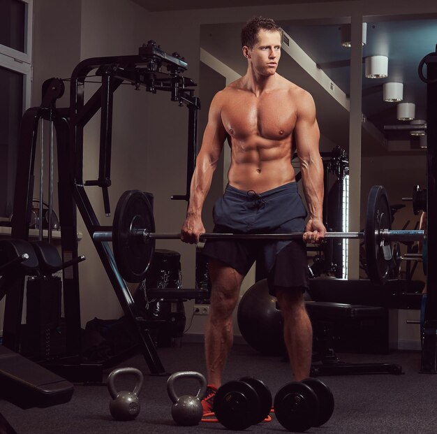 Bodybuilder musclé torse nu faisant un soulevé de terre avec une barre dans une salle de sport.