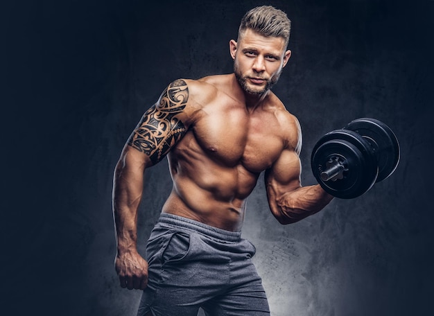 Bodybuilder puissant et élégant avec un tatouage sur son bras, faisant les exercices sur un biceps avec des haltères, regardez la caméra avec un regard confiant. Isolé sur un fond sombre.