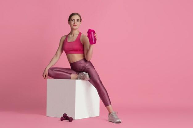 Boire de l'eau. Belle jeune athlète féminine pratiquant, portrait rose monochrome. Entraînement de modèle de coupe sportive avec boîte de saut. Musculation, mode de vie sain, concept de beauté et d'action.