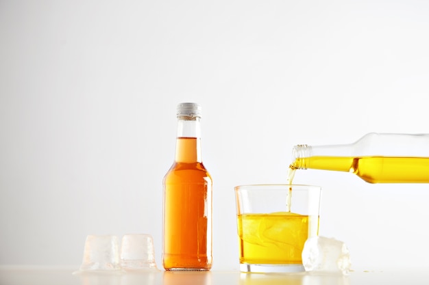 La boisson de limonade savoureuse jaune se déverse de bouteille en verre avec des glaçons près de bouteille fermée scellée non étiquetée avec une boisson orange