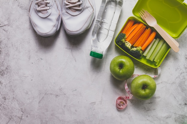 Boîte à lunch avec des légumes à côté de chaussures de sport, bouteille d&#39;eau et des pommes