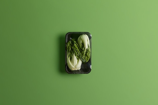 Bok choy ou chou chinois enveloppé d'un film alimentaire sur un plateau noir. Légumes frais à vendre au supermarché isolé sur fond vert. Concept de mode de vie sain, de rafraîchissement et de nutrition