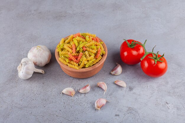 Un bol d'argile de pâtes spirales crues multicolores avec de l'ail et des tomates rouges fraîches.