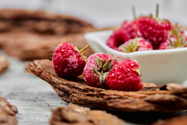 Bol blanc de framboises fraîches avec des morceaux de bois sur une surface grise. Fermer.